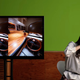 A woman tests the VR headset, while a TV shows the footage of a car she sees inside the headset.